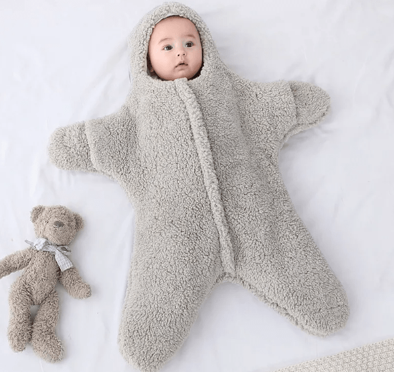 Baby in a fluffy gray onesie lying on a white surface with a teddy bear beside.