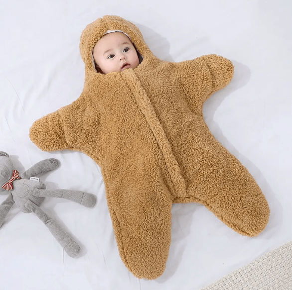 Baby in a brown fleece onesie lying on a white surface with a gray plush toy.