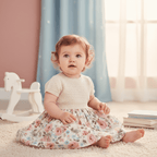 A baby girl sitting on a cream carpet in a nursery, wearing a cream knit short-sleeve top and a matching floral print skirt with pink and blue roses.