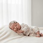 A sleeping newborn baby wearing a matching floral one-piece outfit and bow headband, lying on a soft cream blanket in a bright, softly lit room.